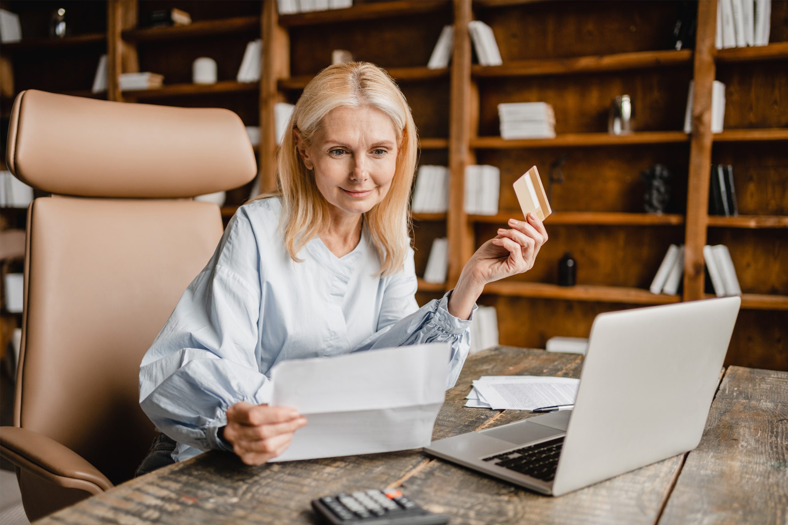 Woman holding mail and credit card with an open laptop at a desk.